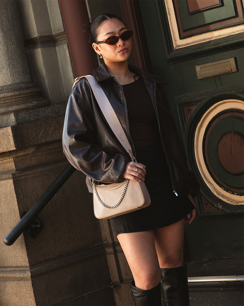 A woman wearing a black leather jacket, black mini skirt, and black knee-high boots, carrying a Jupiter beige shoulder crossbody travel handbag with a silver chain strap. She is standing outdoors in front of a dark green door and stone wall. Travel handbag comes with safety features such as anti-theft zipper, RFID protection and slash proof lining. 