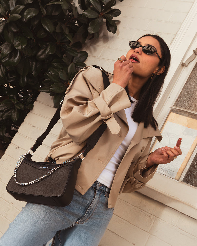 A woman wearing a beige oversized tie-front jacket, white top, blue jeans, and black sunglasses carries a Jupiter  black crossbody travel handbag with a silver chain strap. She is leaning against a white brick wall next to green foliage. Travel handbag comes with safety features such as anti-theft zipper, RFID protection and slash proof lining. 