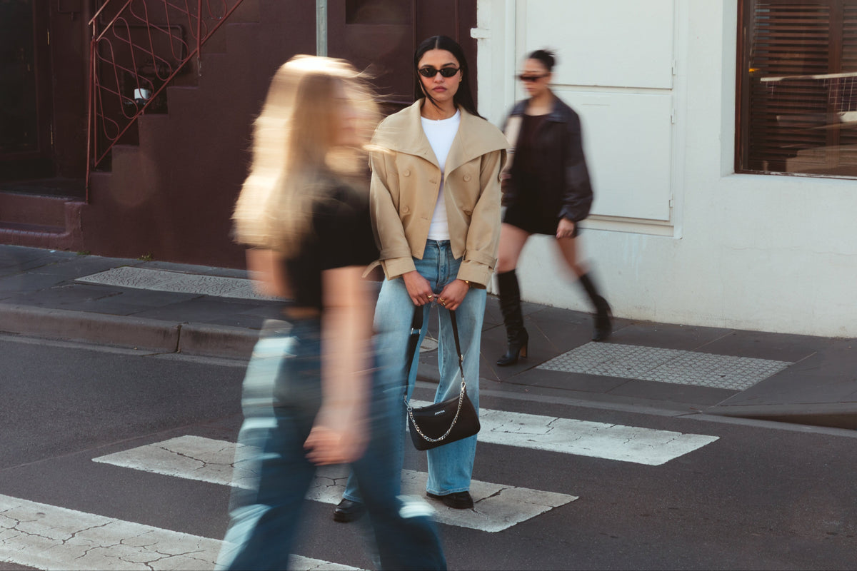A woman stands on a city crosswalk wearing a beige moto jacket, light blue wide-leg jeans, a white top, and sunglasses, holding a Jupiter black crossbody travel handbag. Travel handbag includes safety features such as anti-theft zipper, RFID protection and slash proof lining. 