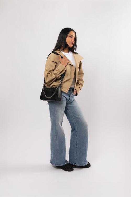A woman modeling a tan utility jacket, light blue wide-leg jeans, a white t-shirt, and a black shoulder travel bag against a plain white studio background. The name Jupiter is branded on the front of the bag. The bag includes safety features to keep valuables safe when travelling overseas or on holidays.