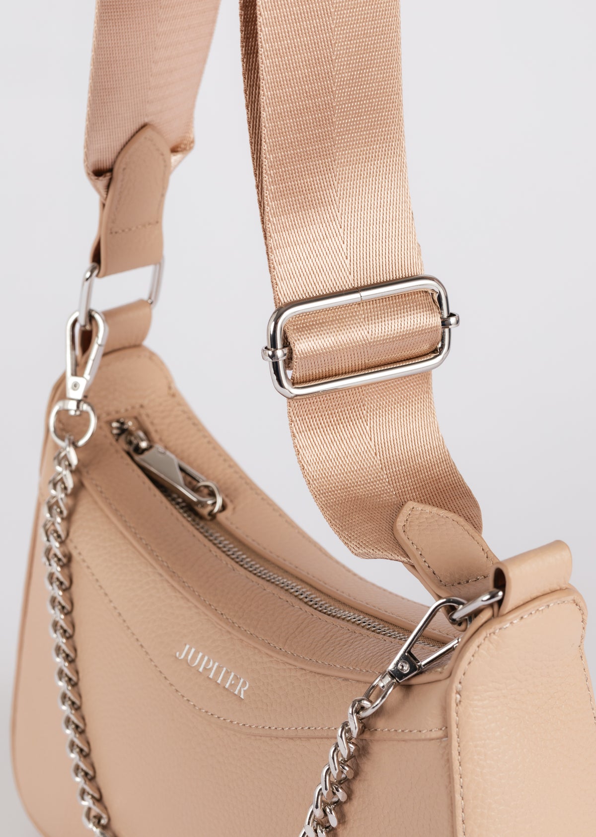Close-up of a Jupiter beige leather shoulder/crossbody travel bag with a matching fabric strap, silver-tone metal hardware, and a chain detail, on a light gray background. 