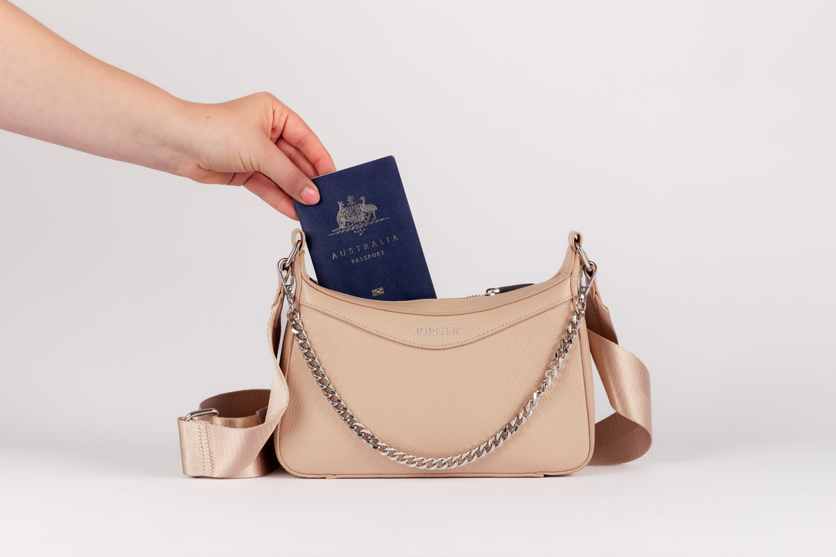 A beige pebbled leather shoulder travel bag with a silver chain strap, shown with a hand inserting a blue Australian passport into its zippered top pocket. The brand name 'JUPITER' is printed in gold on the front of the bag. RFID blocking material keeps valuables like passports safe.
