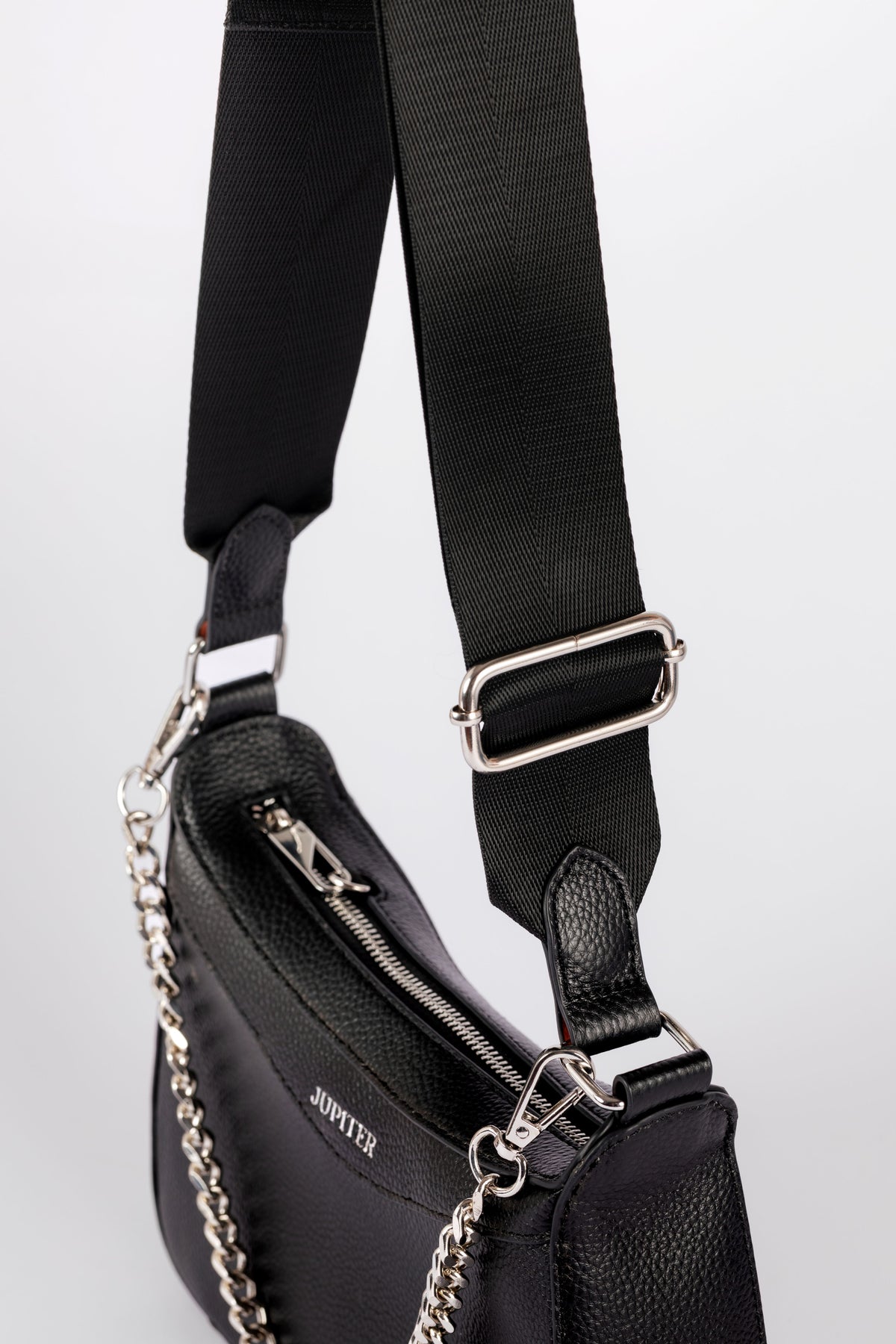 A close-up of a black leather shoulder bag with a wide black webbing strap, silver-tone metal hardware, and a silver chain detail. The brand name 'JUPITER' is visible on the side of the bag. The travel bag includes safety features to keep valuables safe when on holiday or travelling