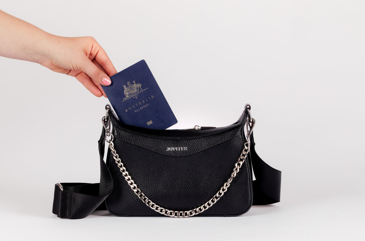 A black pebbled leather shoulder travel bag with a silver chain strap, shown with a hand inserting a blue Australian passport into its zippered top pocket. The brand name 'JUPITER' is printed in gold on the front of the bag. RFID blocking material keeps valuables like passports safe.